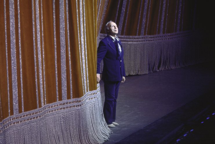 Balanchine bowing in front of the gold curtain in a blue suit and jazz shoes